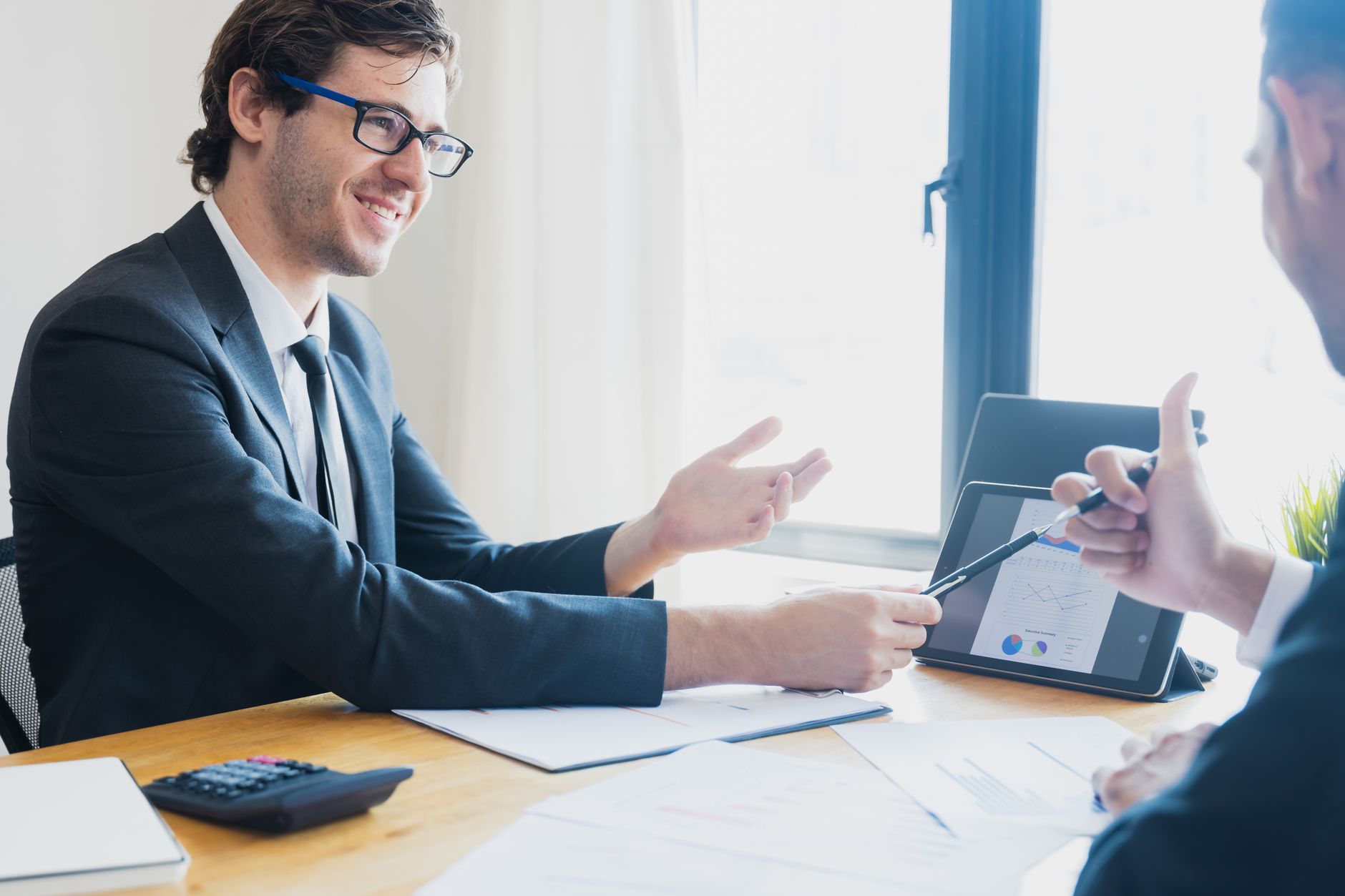 Two businessmen in formal attire discussing a business report in an office setting with a tablet. — How to Trade Foreign Curr