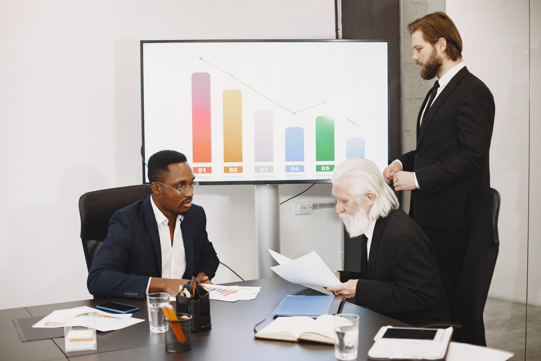 Three businessmen discussing financial data in a modern office setting with charts. — Is the Jeetwin App Right for You? Hones