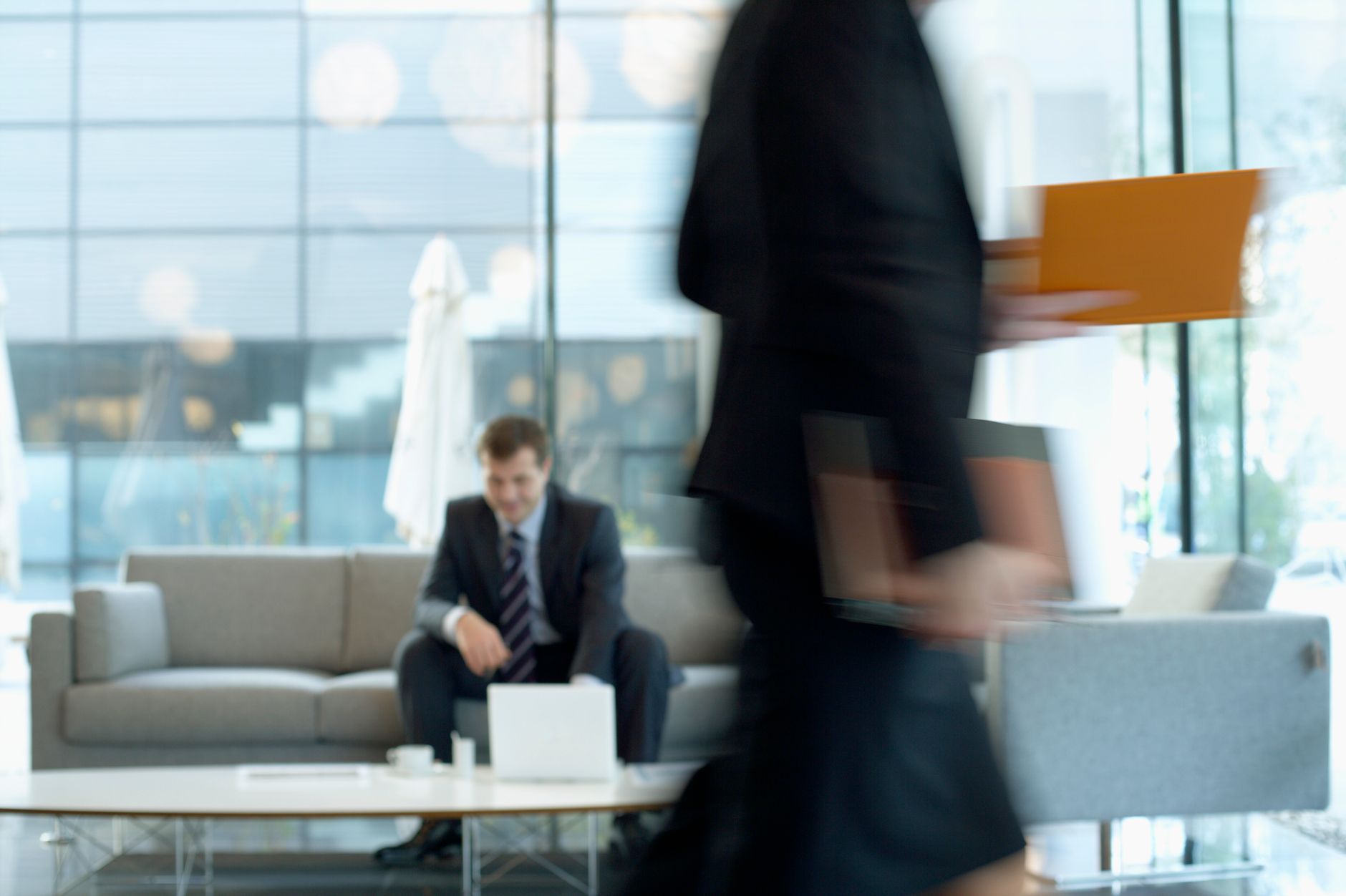 Businessman sitting in modern office lounge using a laptop while colleague walks past, creating dynamic blur. — Mastering the