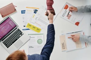 Business professionals engaging in a handshake over a desk filled with financial documents and a laptop. — Play and Earn: Bes