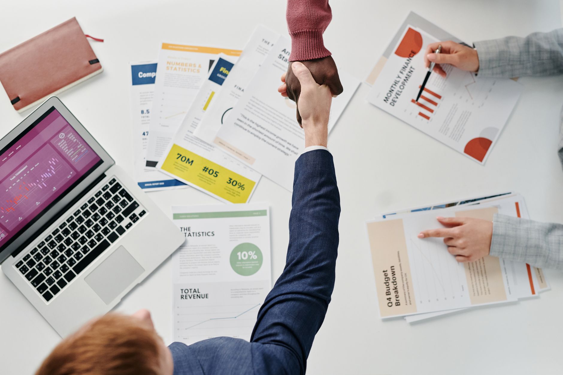 Business professionals engaging in a handshake over a desk filled with financial documents and a laptop. — Play and Earn: Bes