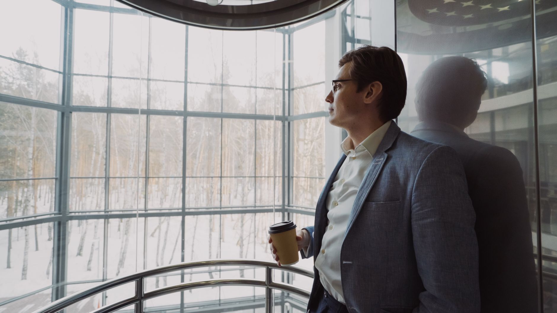 Businessman in blue suit with coffee cup looking out a large window, reflecting on a winter landscape. — Want To Make Your Co
